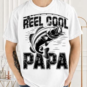 Puede incluir: Camiseta blanca con un gr&aacute;fico negro de un pez saltando fuera del agua, una ca&ntilde;a de pescar y las palabras "REEL COOL PAPA" en una fuente desgastada. La camiseta es de manga corta y tiene cuello redondo.