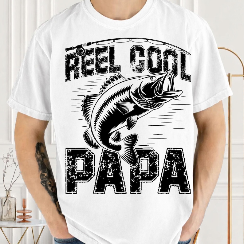 Puede incluir: Camiseta blanca con un gr&aacute;fico negro de un pez saltando fuera del agua, una ca&ntilde;a de pescar y las palabras "REEL COOL PAPA" en una fuente desgastada. La camiseta es de manga corta y tiene cuello redondo.