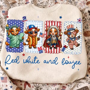 May include: A cream-colored sweatshirt featuring four cartoon cows dressed in patriotic attire. The cows are in rectangular frames with scalloped edges. The text "Red white and boujee" is written in blue script across the bottom.