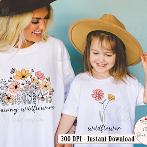 May include: Two white t-shirts with a floral design. The larger shirt has the text "raising wildflowers" and the smaller shirt has the text "wildflower".