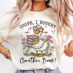 Könnte beinhalten: Cremefarbenes Sweatshirt mit einer skurrilen Grafik einer Ente mit herzförmiger Brille, die Bücher hält. Der Text lautet "OOOPS, I BOUGHT Another Book!" umgeben von rosa Blumen und Sternen. Ein lustiges, buchbezogenes Design.