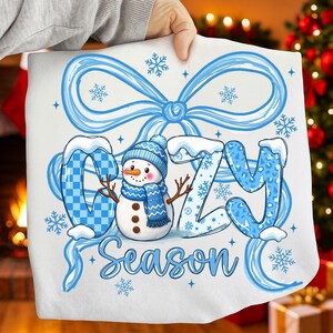May include: A white pillow with a blue bow and snowflakes. The word "COZY" is written in blue, with the letter "O" in a blue and white checkered pattern. A snowman with a blue hat and scarf is in the center. The word "Season" is written below.