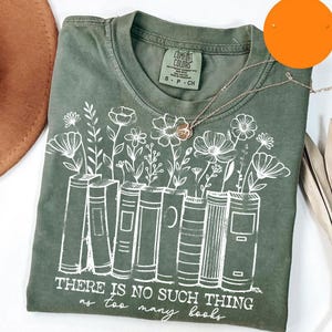 Puede incluir: Camiseta verde oliva con un gr&aacute;fico blanco de libros con flores que crecen. El texto "THERE IS NO SUCH THING as too many books" est&aacute; impreso debajo. Un collar plateado con un colgante de flor est&aacute; sobre la camiseta.