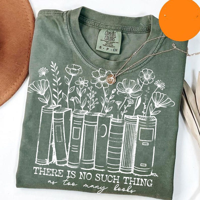 Puede incluir: Camiseta verde oliva con un gr&aacute;fico blanco de libros con flores que crecen. El texto "THERE IS NO SUCH THING as too many books" est&aacute; impreso debajo. Un collar plateado con un colgante de flor est&aacute; sobre la camiseta.