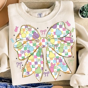 May include: Cream-coloured sweatshirt with a large bow design outlined in gold. The bow features a checkered pattern and colourful Easter-themed graphics, including bunnies, chicks, and bows. Small bunny and bow graphics are scattered around the bow.