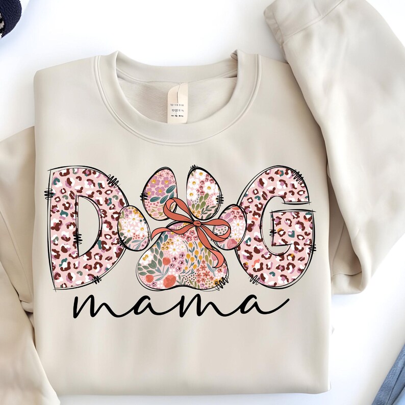 May include: A beige sweatshirt with the words "Dog Mama" printed on the front. The letters are outlined in black and filled with a pink and brown leopard print. A paw print with a pink bow is in the centre of the word "Dog".