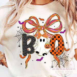 May include: Cream-colored Halloween-themed t-shirt with the word "BOO" in a playful design. The "B" is black with a leopard print, the "O" is orange with black polka dots, and the second "O" is a ghost holding a pumpkin.