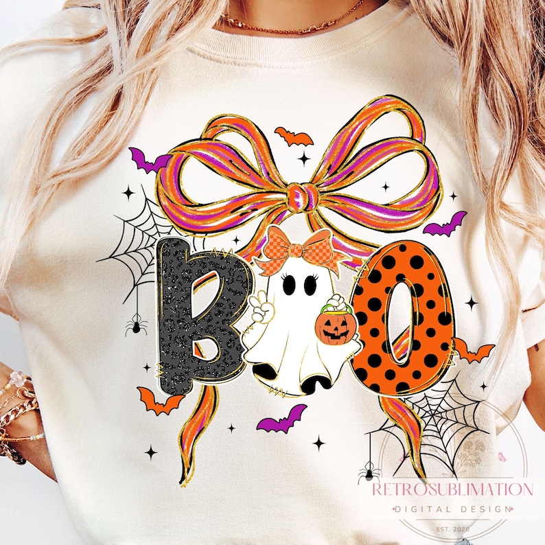 May include: Cream-colored Halloween-themed t-shirt with the word "BOO" in a playful design. The "B" is black with a leopard print, the "O" is orange with black polka dots, and the second "O" is a ghost holding a pumpkin.