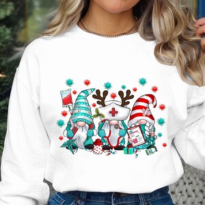 Puede incluir: Sudadera blanca con un gr&aacute;fico de tres gnomos vestidos de enfermeras. Los gnomos llevan gorros rojos y blancos, camisas a rayas azules y blancas y batas blancas. Est&aacute;n sosteniendo suministros m&eacute;dicos, incluyendo un estetoscopio, una jeringa y un portapapeles. El fondo es blanco con un patr&oacute;n de copos de nieve verdes y rojos.
