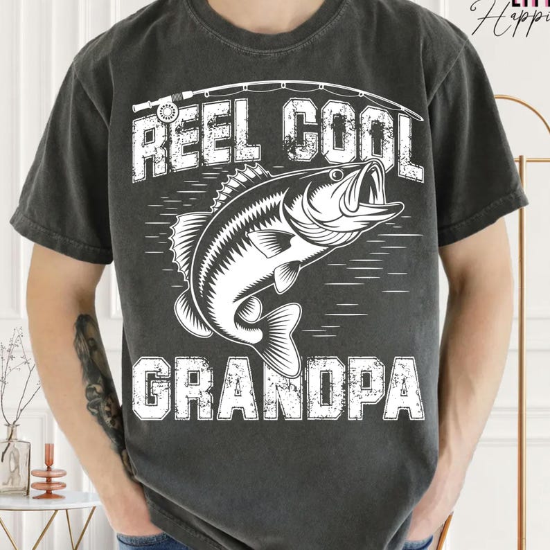 Puede incluir: Camiseta gris oscuro con un gr&aacute;fico blanco. El gr&aacute;fico presenta un pez y las palabras "REEL COOL GRANDPA". La camiseta tiene mangas cortas y cuello redondo. El dise&ntilde;o est&aacute; centrado en la parte delantera de la camiseta.