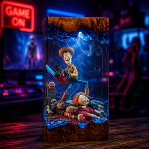 Op de afbeelding: Een rechthoekige harslamp met een houten voet en bovenkant, met daarin een miniatuurscène van een cowboyfiguur met een kettingzaag, staand over een gevallen pop en een skelet bij een kleine grafsteen, verlicht door blauw intern licht.