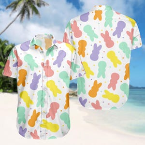 May include: White short-sleeved button-down shirt with a colorful pattern of cartoon bunny shapes in orange, yellow, purple, pink, and green. The shirt is displayed on a beach background, showing the front and back.