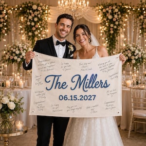 May include: A rectangular off-white fabric wedding sign featuring the blue embroidered text The Millers and the date 06.15.2027. The banner includes various handwritten-style congratulatory messages from guests and has metal grommets in each corner.