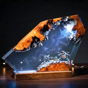 Peut inclure: Une lampe décorative composée d'une tranche de bois naturel enchâssée dans de la résine transparente aux bords irréguliers. Trois petites figurines bleues et grises reposent sur une base en bois enneigée. Une lumière émet des volutes de fumée.