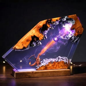 May include: A rectangular resin and wood lamp featuring a diorama of a dragon and a psychic creature. The dragon breathes a stream of orange smoke toward a purple glowing orb held by the creature, set within a clear resin block with wood edges.