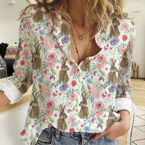 May include: A white button-down shirt with a floral and bunny print. The long-sleeved shirt features a V-neck and is decorated with pink, purple, and blue flowers, along with brown bunnies. The cuffs are rolled up.