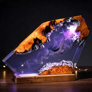 Op de afbeelding: Een decoratieve lamp met een frame van gepolijst hout rondom een blok heldere hars. Binnenin verlicht een paarsachtig licht een kleine, gevleugelde drakenfiguur omringd door rookpluimen, geplaatst tegen een grillige, natuurlijke houten rand.