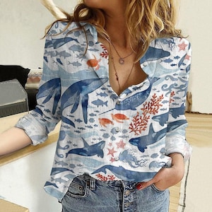 May include: A long-sleeved button-down shirt with a marine life design. The shirt has a light blue and white background, with illustrations of whales, fish, coral, and jellyfish. It features a classic collar and button closure.