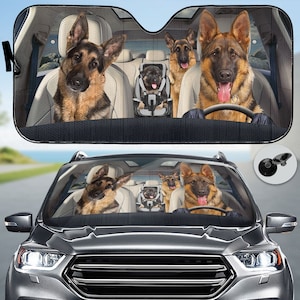 Op de afbeelding: Auto zonnescherm met een humoristisch ontwerp van Duitse herders in een auto. De honden worden afgebeeld op de bestuurdersstoel, de passagiersstoel en een kinderzitje. Het zonnescherm is zwart met een achtergrond van een auto-interieur.