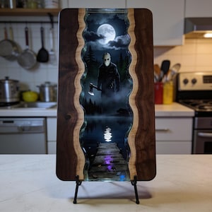 May include: A rectangular live-edge wood serving board featuring a central resin inlay depicting a night scene with a hockey-masked figure holding an axe, a full moon, a forest, a wooden dock, and a lake reflection in dark blue and black tones.