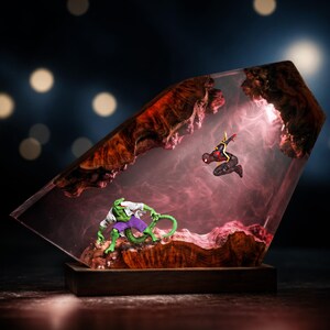 May include: A decorative resin and wood lamp featuring a miniature green lizard-like figure in a white lab coat and purple shorts facing a black and red superhero figure suspended in a translucent, smoky pink-tinted resin block on a base.