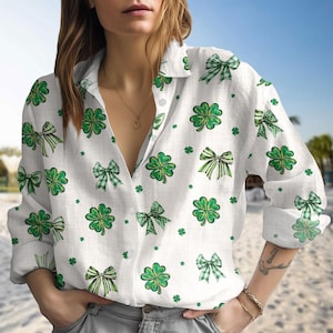 May include: White button-down shirt featuring a pattern of green shamrocks and bows. The bows have solid and plaid designs. The shirt has long sleeves and a collared neckline. The shirt is made of a lightweight fabric.