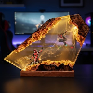 May include: A decorative lamp featuring a clear resin block with embedded wood edges, illuminated from within. Inside the resin are two miniature action figures: a red and black Deadpool character in a kneeling pose and a leaping Spider-Man figure.