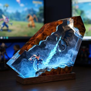 May include: A decorative resin and wood lamp featuring a miniature Greninja and Charizard battling within a clear, smoke-filled block. The scene depicts a blue energy beam connecting the two figures, set on a rectangular wooden base.