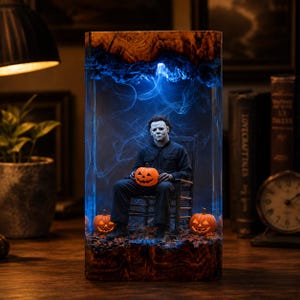 May include: A rectangular resin lamp featuring a dark wood base and top, enclosing a miniature figure of a masked character in a dark jumpsuit sitting on a wooden chair. The scene includes three orange jack-o'-lanterns and glowing blue smoke.