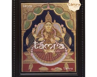 May include: A framed painting of a Hindu goddess in a red and gold robe, sitting on a throne with a lotus flower in her lap. The painting is surrounded by a black frame with gold accents. The painting is titled "tamora handicrafts".