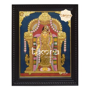 May include: A framed painting of a Hindu deity in a golden throne with a blue background. The deity is adorned with gold jewelry and flowers. The painting is framed in black wood with a gold trim.