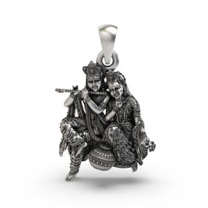 Puede incluir: Un colgante plateado que representa a dos figuras, una de las cuales toca la flauta. Las figuras están adornadas con joyas intrincadas y sentadas cerca de una olla decorativa. El colgante tiene un lazo para una cadena.