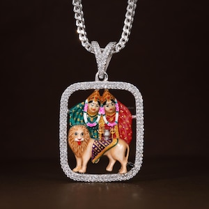 May include: A silver pendant necklace featuring a rectangular pendant with a detailed, colorful depiction of two figures and a lion. The pendant is framed with sparkling stones and hangs from a silver chain.