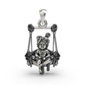 Könnte beinhalten: Ein silberner Anhänger mit einer detaillierten Darstellung einer sitzenden Figur auf einer Schaukel. Die Figur trägt eine dekorative Kopfbedeckung mit einer Feder und ist mit Schmuck geschmückt. Die Schaukel ist mit floralen Akzenten verziert und hängt an einer gebogenen Stange.