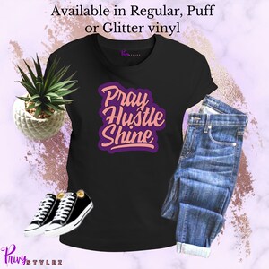 May include: Black t-shirt with the text "Pray Hustle Shine" in pink and purple lettering. The shirt is available in regular, puff, or glitter vinyl.