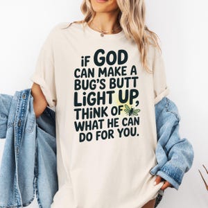 May include: Cream-colored t-shirt with the text "If God can make a bug's butt light up, think of what he can do for you." The design includes a stylized firefly illustration. The shirt is a casual, comfortable style.