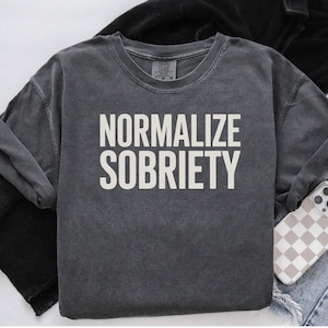 May include: A charcoal gray sweatshirt with the words "NORMALIZE SOBRIETY" printed in white block letters. The long-sleeved shirt is made of a soft material and has a crew neck. A phone with a checkered case rests on the side.