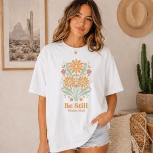 Może przedstawiać: Biały t-shirt z krótkim rękawem z centralną grafiką przedstawiającą stylizowane kwiaty w odcieniach pomarańczu, żółci i szałwiowej zieleni. Pod kwiatowym wzorem znajduje się napis "Be Still" pogrubioną czcionką oraz "Psalm 46:10" mniejszym drukiem.