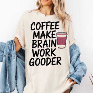 Peut inclure: T-shirt crème avec l'inscription noire "COFFEE MAKE BRAIN WORK GOODER". Un dessin de tasse à café rose et blanche est à droite du texte. Le t-shirt a des manches retroussées et est porté avec une veste en jean.