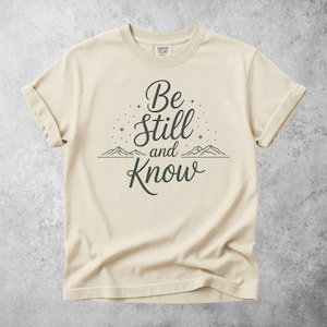 Puede incluir: Camiseta color crema con mangas enrolladas. La camiseta presenta el texto "Be Still and Know" en una escritura gris oscuro, con un gráfico de cordillera debajo. La camiseta está hecha de un material suave.