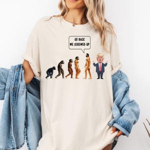 Puede incluir: Camiseta color crema con un gráfico de evolución. El gráfico representa la evolución del hombre, culminando con una caricatura de un hombre con traje y el texto "GO BACK WE SCREWED UP" en un bocadillo.