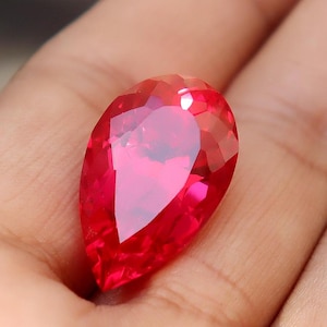 Può includere: Una gemma rossa sfaccettata a forma di pera. La gemma è tenuta in una mano, mostrando il suo taglio brillante e il suo colore vibrante. Le sfaccettature della pietra riflettono la luce, creando un effetto scintillante. La forma è allungata.