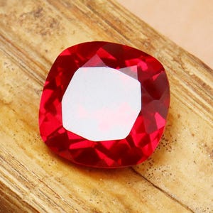 Könnte beinhalten: Ein leuchtender, kissenförmig geschliffener roter Edelstein mit facettierter Oberfläche, die das Licht reflektiert. Der Stein hat einen tiefen, gesättigten karmesinroten Farbton und eine symmetrische, abgerundete quadratische Form.