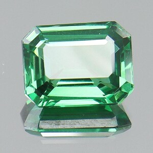May include: A rectangular-cut, emerald-green gemstone with a reflective surface. The gem is faceted, showcasing a bright, light-green interior. The gemstone is displayed on a reflective surface, creating a mirrored effect.
