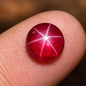 May include: A round, cabochon-cut star ruby gemstone featuring a deep, vibrant red color and a distinct, centered six-rayed star pattern that reflects light across its smooth, polished surface, showcasing the natural asterism of the stone.