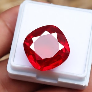 Puede incluir: Un vibrante rubí tallado en cojín, exhibido en una caja de plástico blanco. Las facetas del rubí reflejan la luz, mostrando su color rojo intenso y brillo interno. La gema tiene forma cuadrada con esquinas redondeadas.