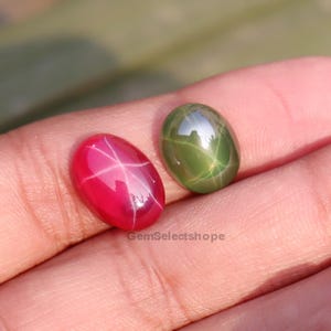 Op de afbeelding: Twee ovale edelstenen. Eén is een levendig rood met een zesstralig sterpatroon, de andere is een groene edelsteen met een vergelijkbaar stereffect. De edelstenen zijn glad en gepolijst en reflecteren licht. De foto is gemaakt in natuurlijk licht.