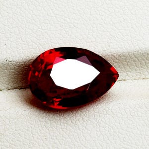 Pode incluir: Uma pedra preciosa facetada em forma de pêra, com um tom vermelho profundo. A gema tem um grande centro branco e reflexivo e está colocada sobre uma superfície texturizada esbranquiçada. O corte da pedra cria múltiplas facetas.