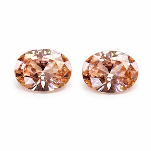 May include: Two oval-cut gemstones with a light peach and champagne color. Each stone features a faceted surface that reflects light, creating a brilliant, sparkling appearance with multiple geometric patterns across the top of the stones.