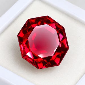Op de afbeelding: Een levendige, dieprode edelsteen geslepen in een nauwkeurige achthoekige vorm met meerdere facetten die het licht reflecteren. De steen is geplaatst tegen een effen, lichtgekleurde achtergrond, wat de intense kleurverzadiging benadrukt.